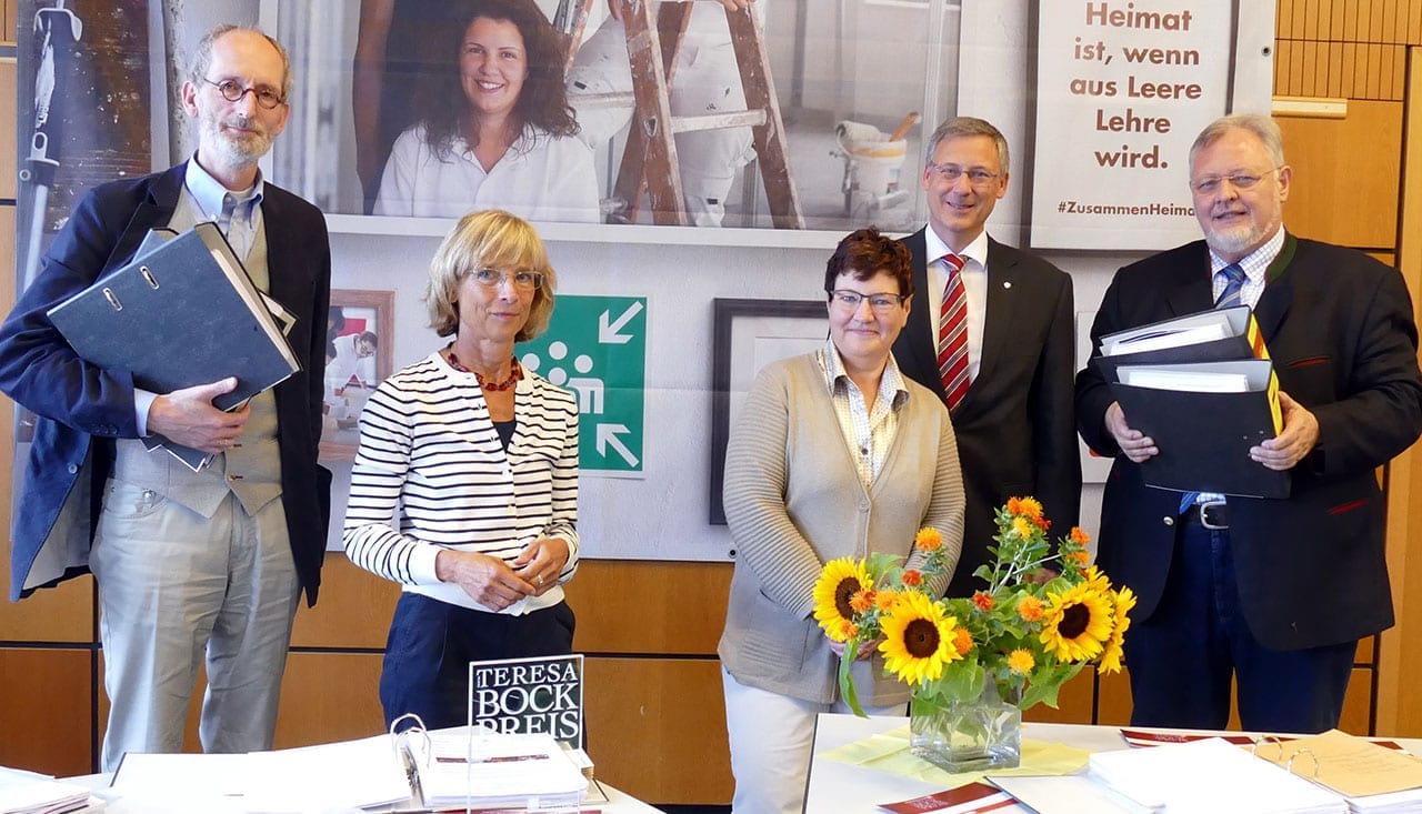 Fünf Erwachsene stehen hinter einem Tisch mit Sonnenblumen, Büchern und Ordnern und posieren für ein Gruppenfoto bei einer Indoor-Veranstaltung. Im Hintergrund ist ein großes Plakat mit einer lächelnden Frau und deutschem Text zu sehen.